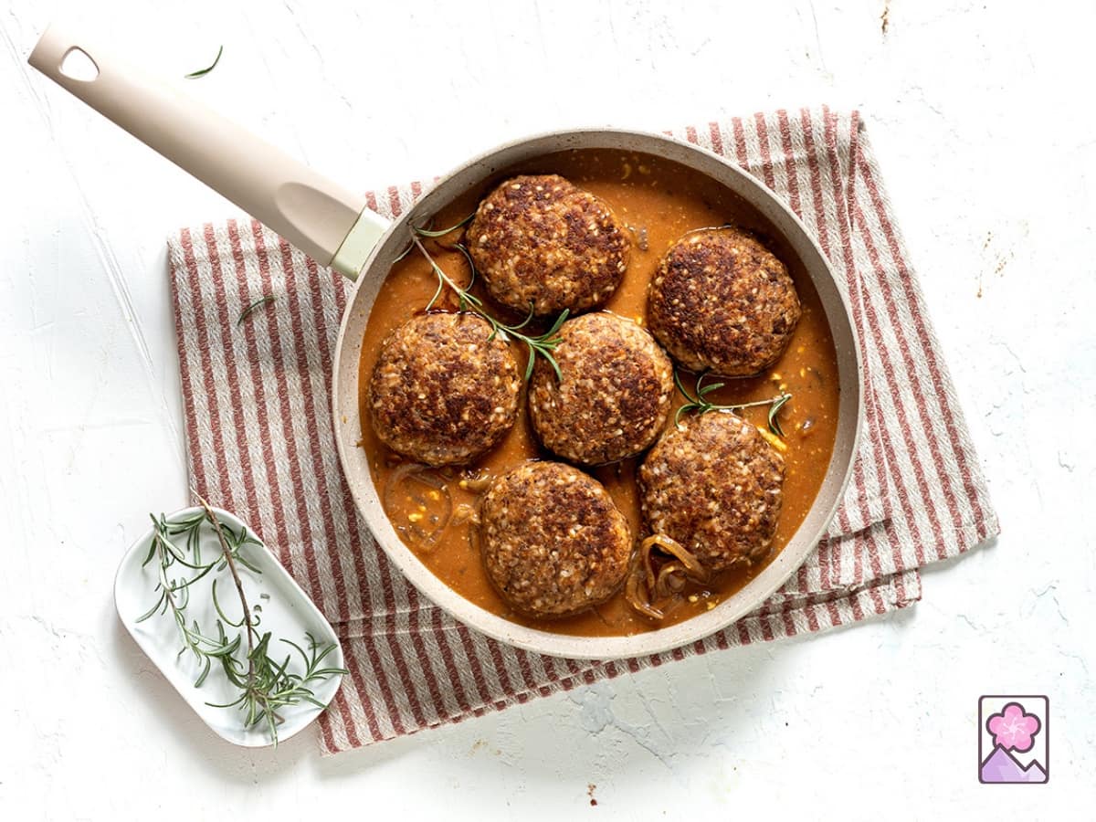 Six Salisbury steaks simmering in sauce in a pan topped with herbs.