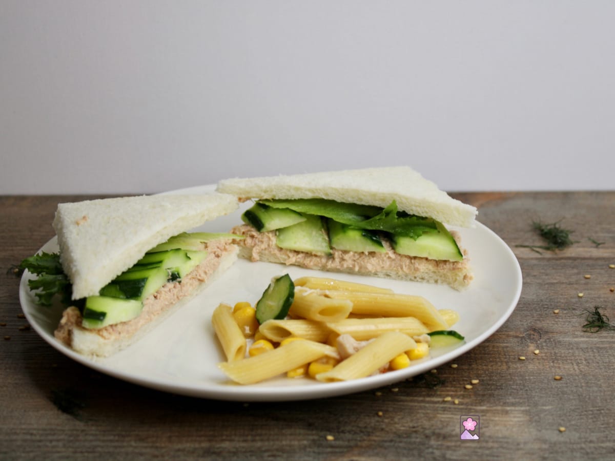 A simple Japanese tuna salad sandwich on white bread. The crust is trimmed off and ithe sandwich is filled with tuna salad, cucumber and lettuce. It s served on a white plate with a side of pasta salad.