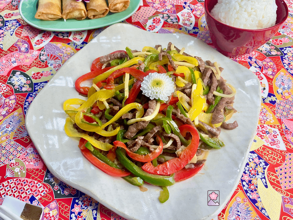 Stir fried pepper steak on a white serving tray with egg rolls and rice in the background