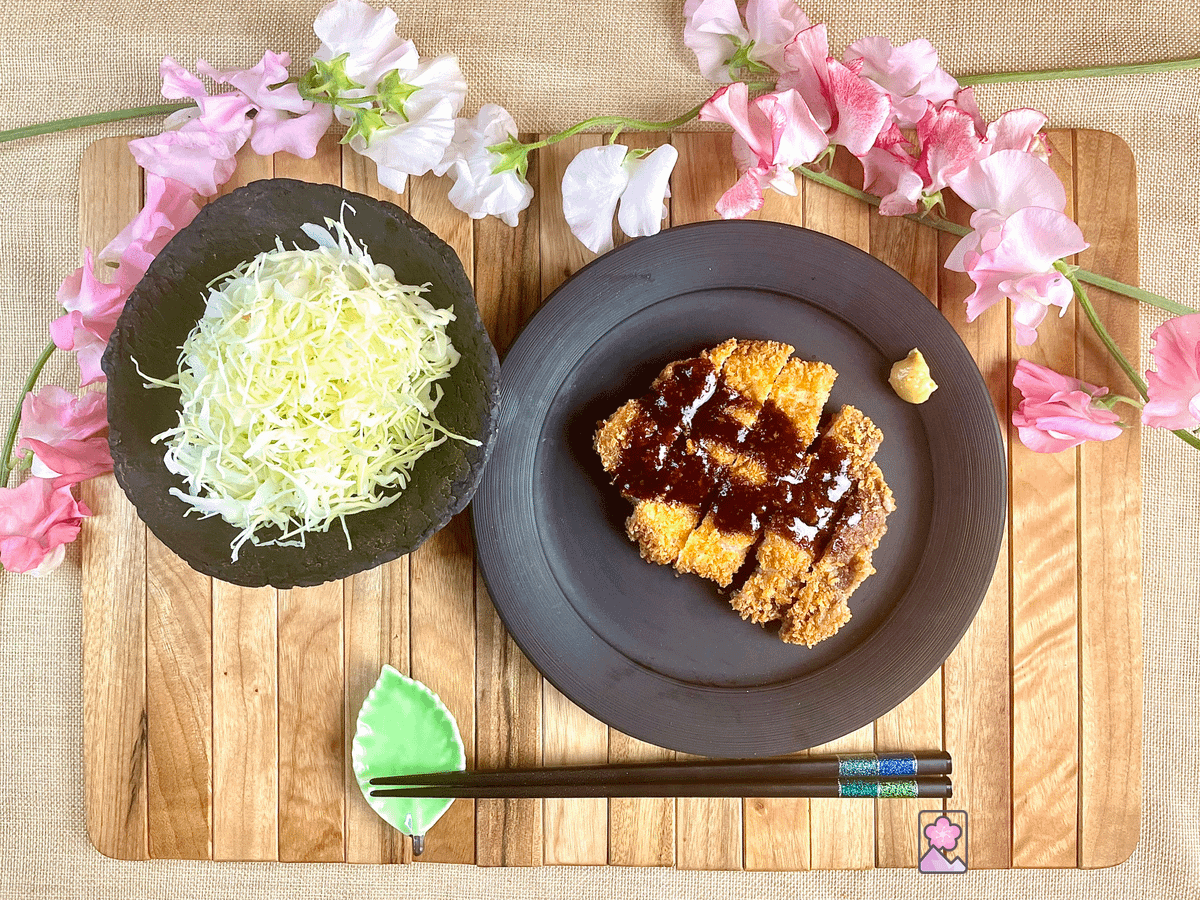 A tonkatsu fried pork cutlet on a black plate topped with brown sauce and some yellow mustard. Behind it is a small black bowl of shredded cabbage