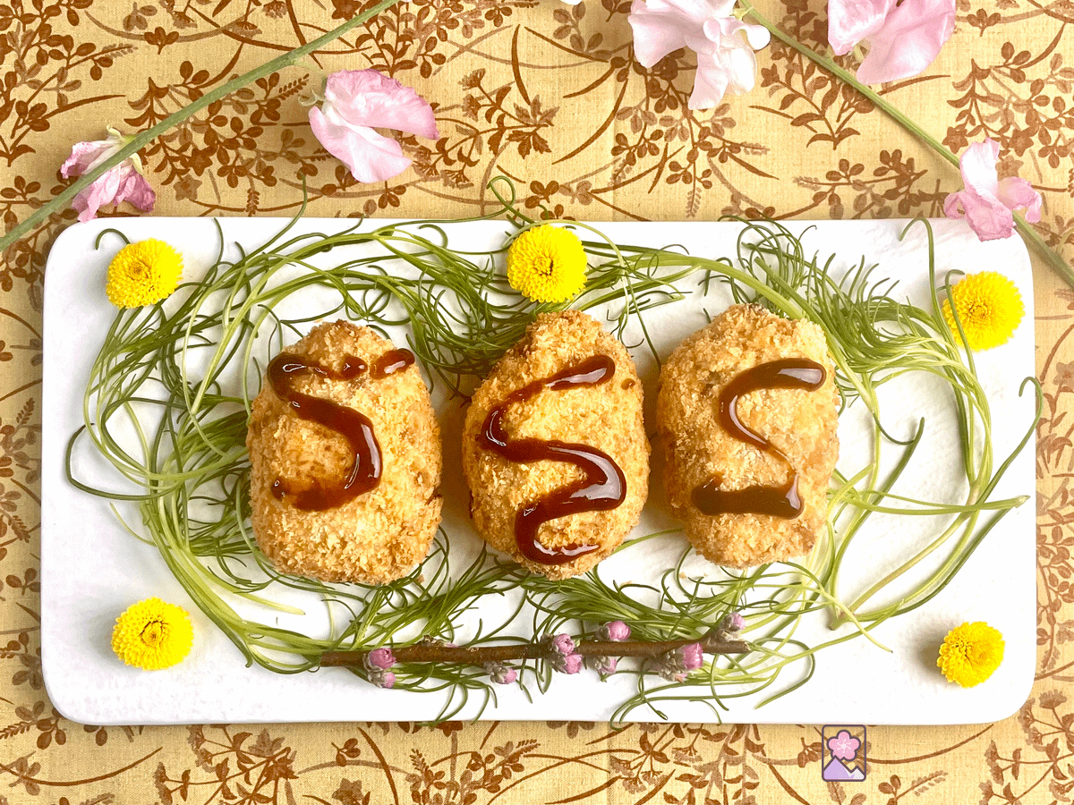 3 croquettes on a white plate sitting on top of some greens and surrounded by flowers. The croquettes have brown sauce on them.