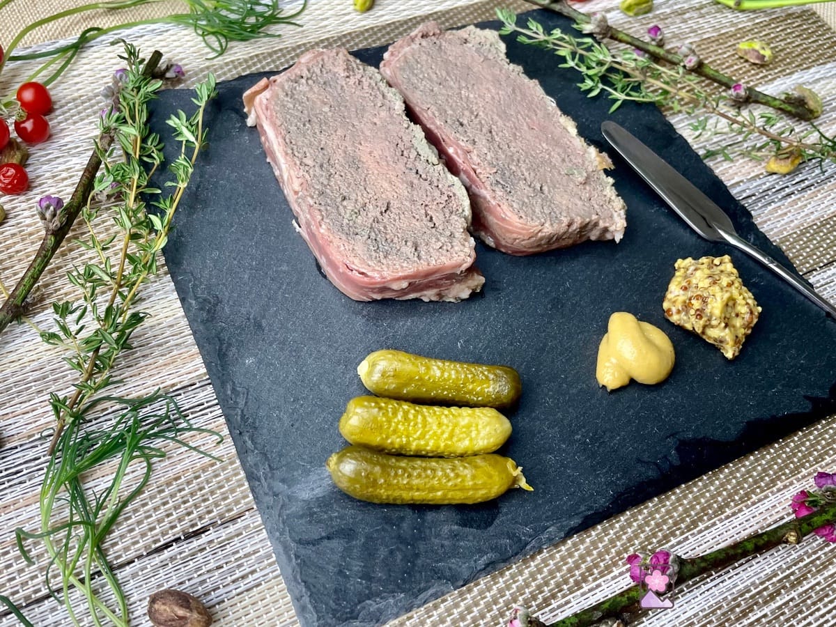Two pieces of pate on a black slate type with pickles and 2 type of mustard.
