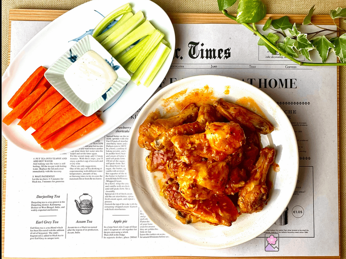 Baked crispy chicken wings slathered in sweet and spicy buffalo chicken sauce. Served on a white plate. Behind it there are celery and carrot sticks and a dish of ranch dressing.