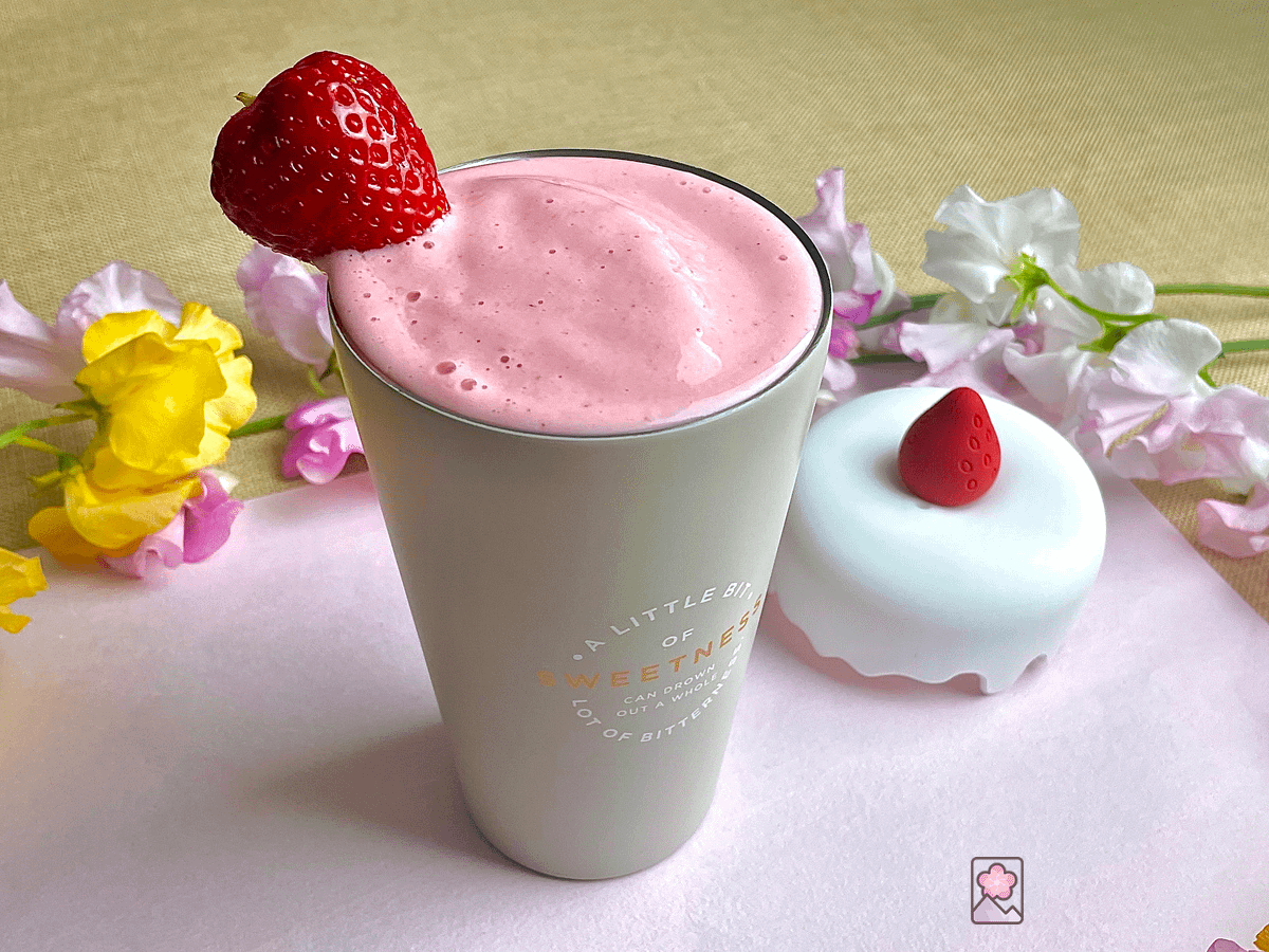 A beautiful pink smoothie in a tall metal cup with a strawberry on the rim. The cup lid is next to the drink and flowers are in the background.