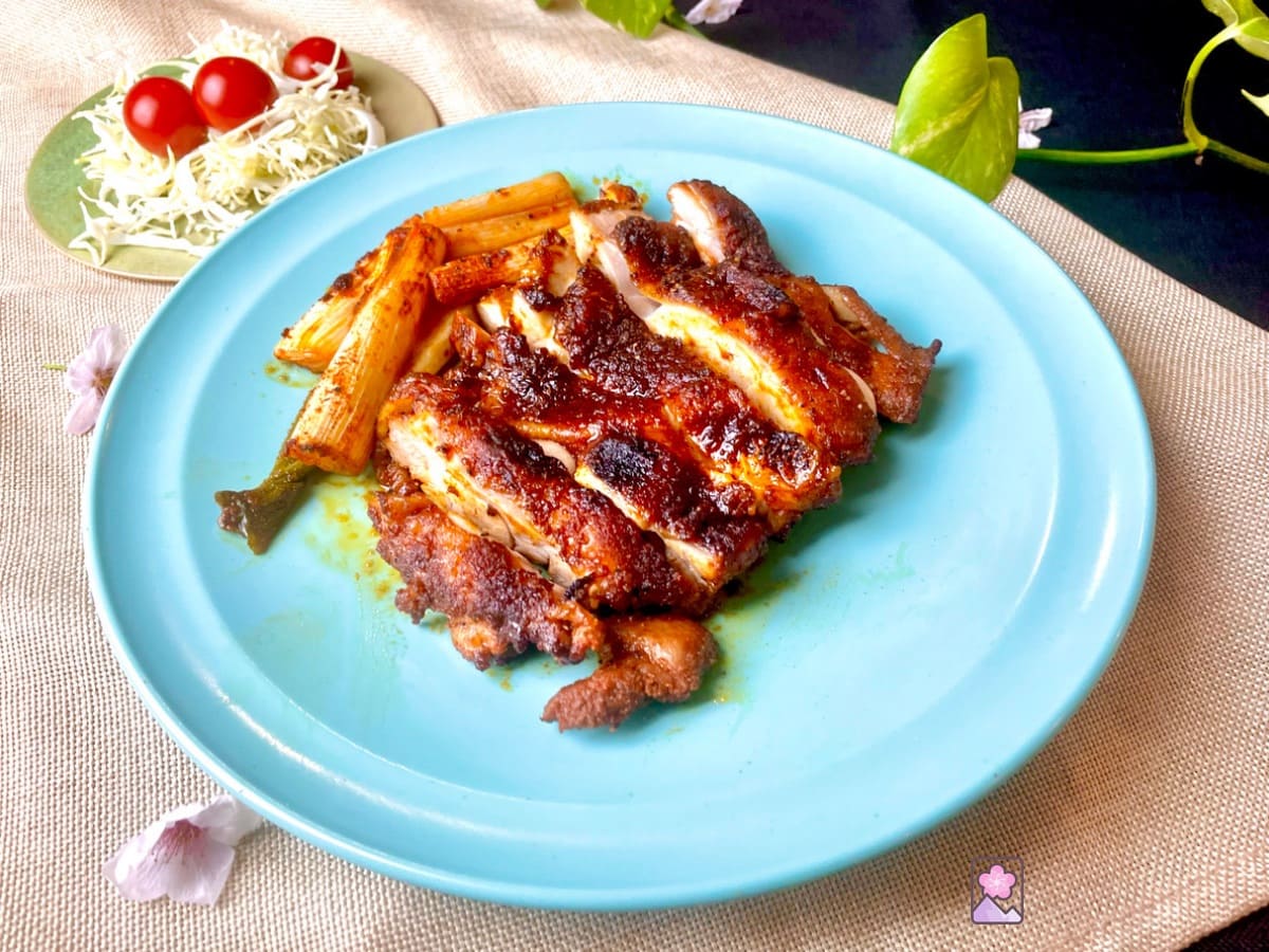 A large chicken thigh coated with red barbecue spices and sliced evenly. There are leeks on the same plate. Behind it there is a cabbage salad with cherry tomatoes on top.