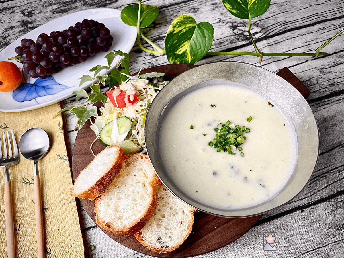 Cold potato soup sprinkled with chopped chives is served in a silver bowl on a wooden board. Sliced baguette and a fresh salad are next to the soup,
