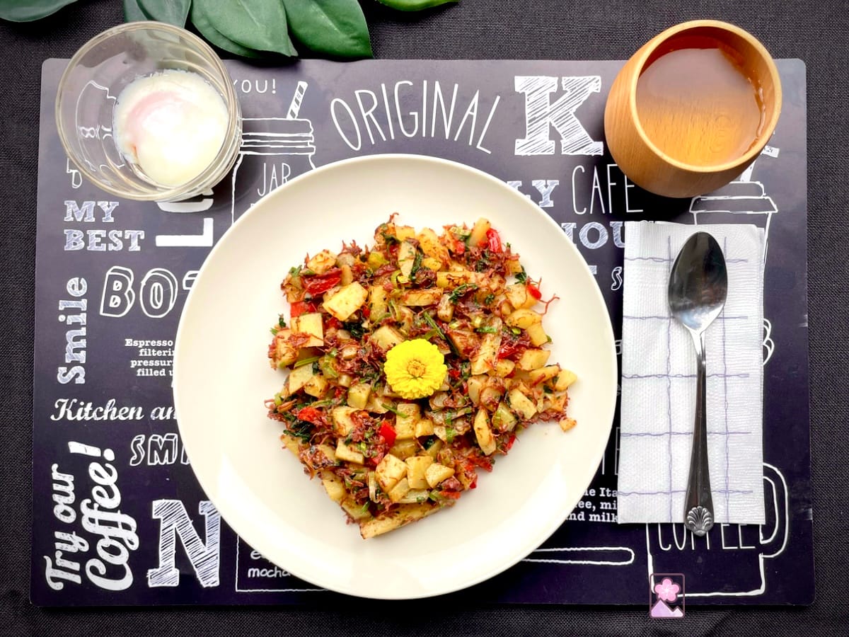 Savory Corned beef hash on a white plate topped with a dandelion flower. There is a poached egg in a glass to the top left and a wooden cup with tea to the top right.