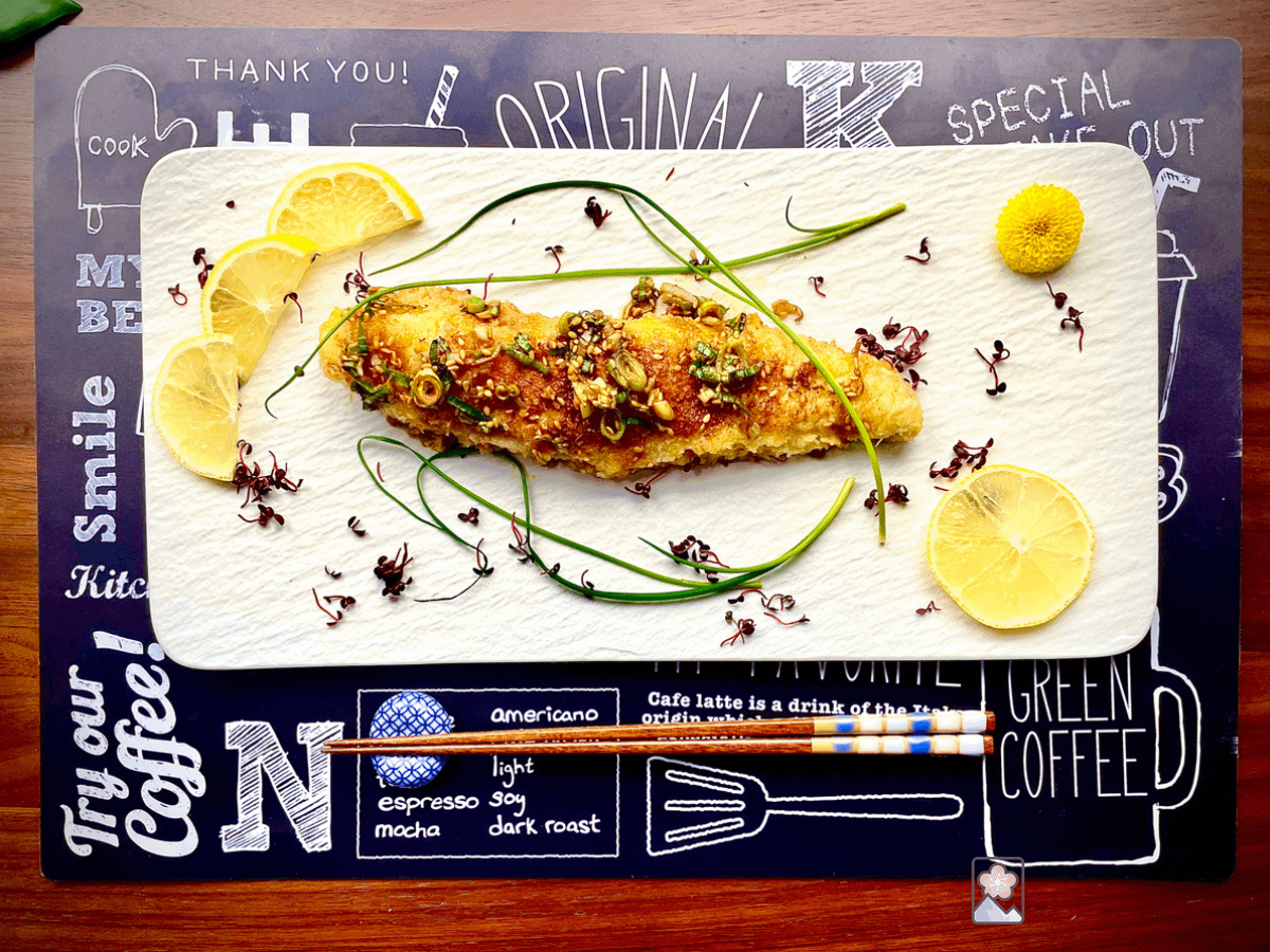 A delicious piece of fried fish on a rectangular white plate. The fish has lemon slices behind it and is topped with onion grass. The plate is on a black placemat with a cafe menu print.