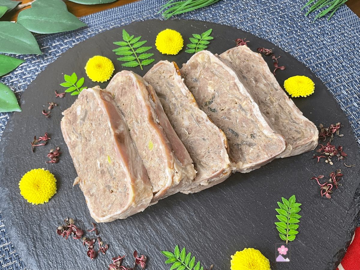 5 rectangular slices of prime rib terrine wrapped in bacon. They are on a slate plate with leaves and flowers arranged decoratively around the terrine.