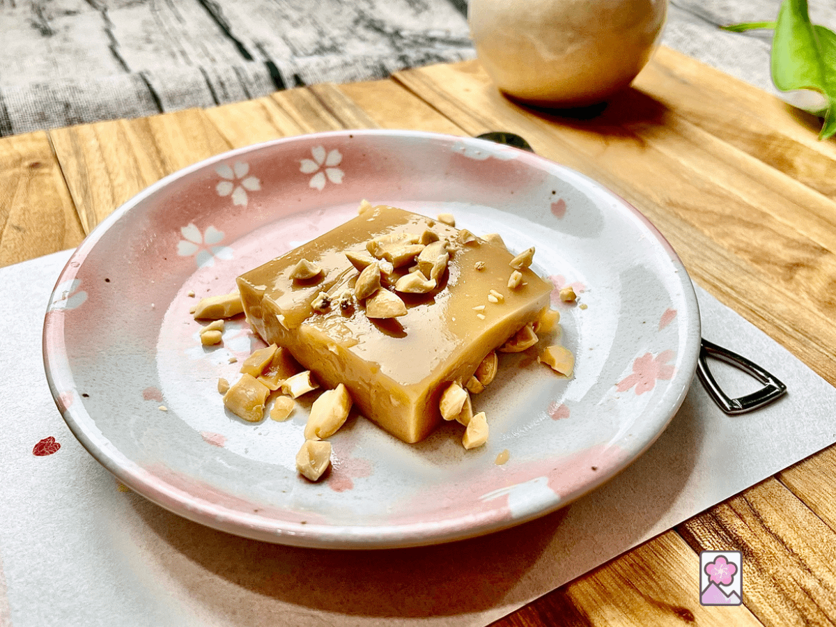 A creamy square piece of peanut tofu on a small ceramic plate on a wooden placemat. There are crushed peanuts sprinkled on top of the tofu.