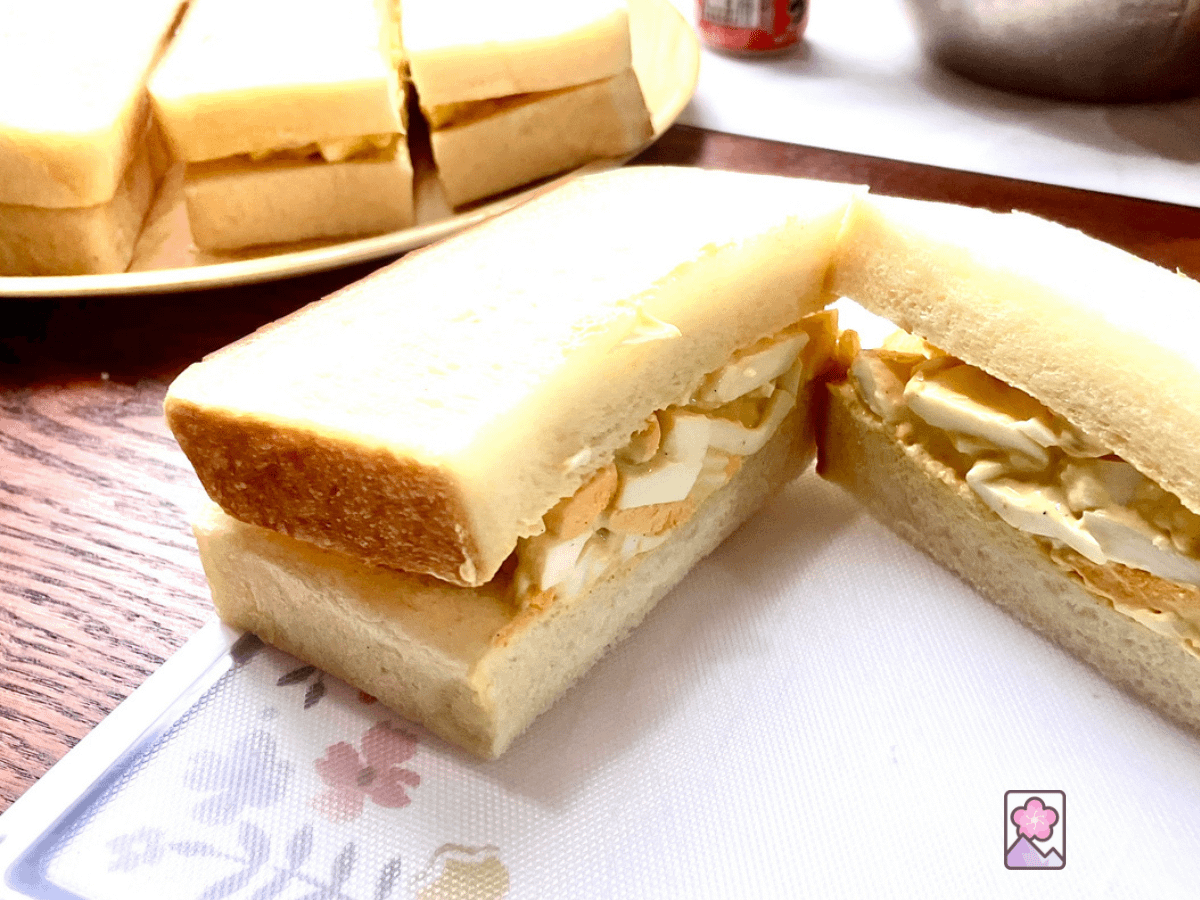 A Japanese Egg Salad Sandwich on a cutting board cut in half lengthways. There are 3 half sandwiches on a tray in the background