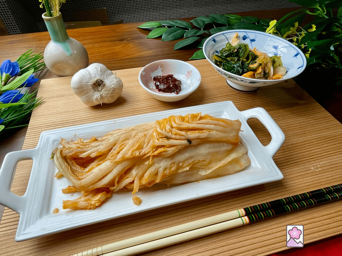 Japanese Style Kimchi Is served on a long flat plate where slices of Napa kimchi are piled. Just behind it there is a bowl with leafy pieces of napa. It is served on a grooved wooden tray. Style Kimch