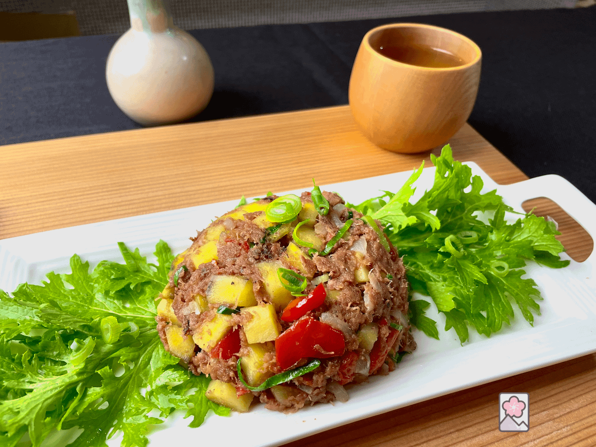 Corned Beef Hash with Japanese Sweet Potato is scooped onto a bed of wasabi greens on a rectangular plate. It’s served on a wooden tray with a wood cup of tea in the background and a small flower vase.