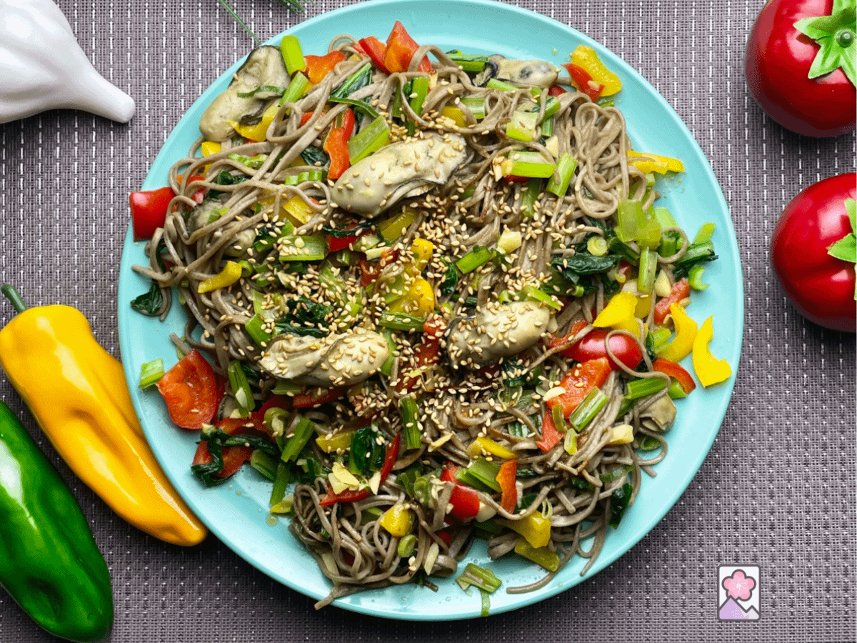 Spicy oyster yakisoba topped with sesame seeds on a plate surrounded by to,stores, garlic and peppers.