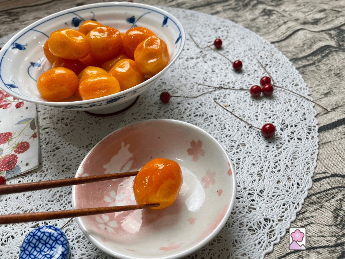 Poached Kinkan with a single Kinkan on a plate is picked up with chopsticks while a bowl of Kinkan sits in the background