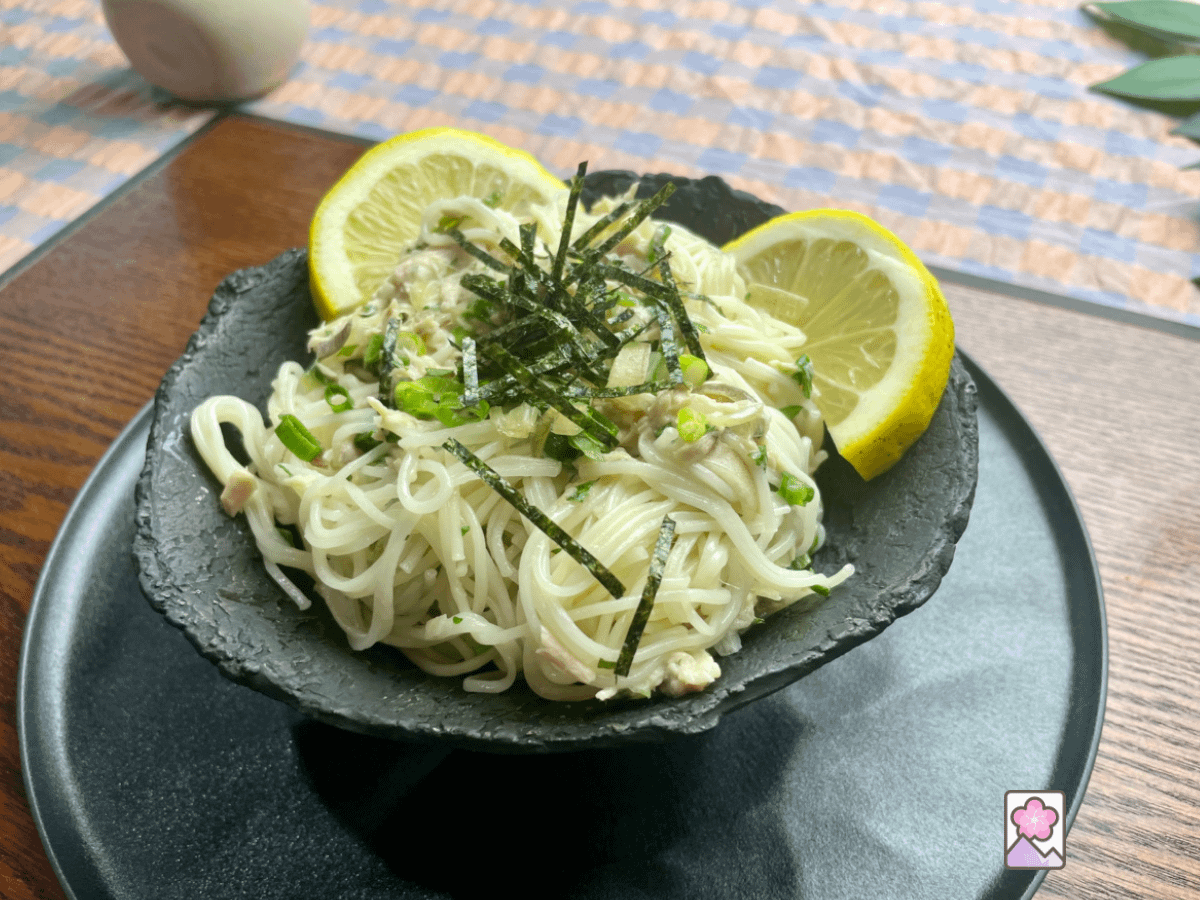 Tuna Somen features long thin some. Noodles in a black ceramic bowl on a resin plate. It’s topped with seaweed strips and lemon wedges.