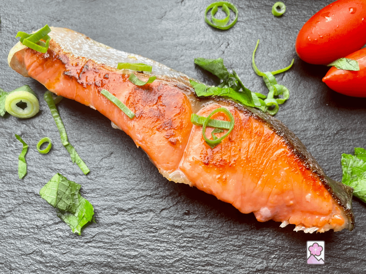 A think sliced filet of Salmon that was marinated in Koji on a plate sprinkled with fresh green herbs and some cherry tomatoes in the background. The fish is on a slate tray.