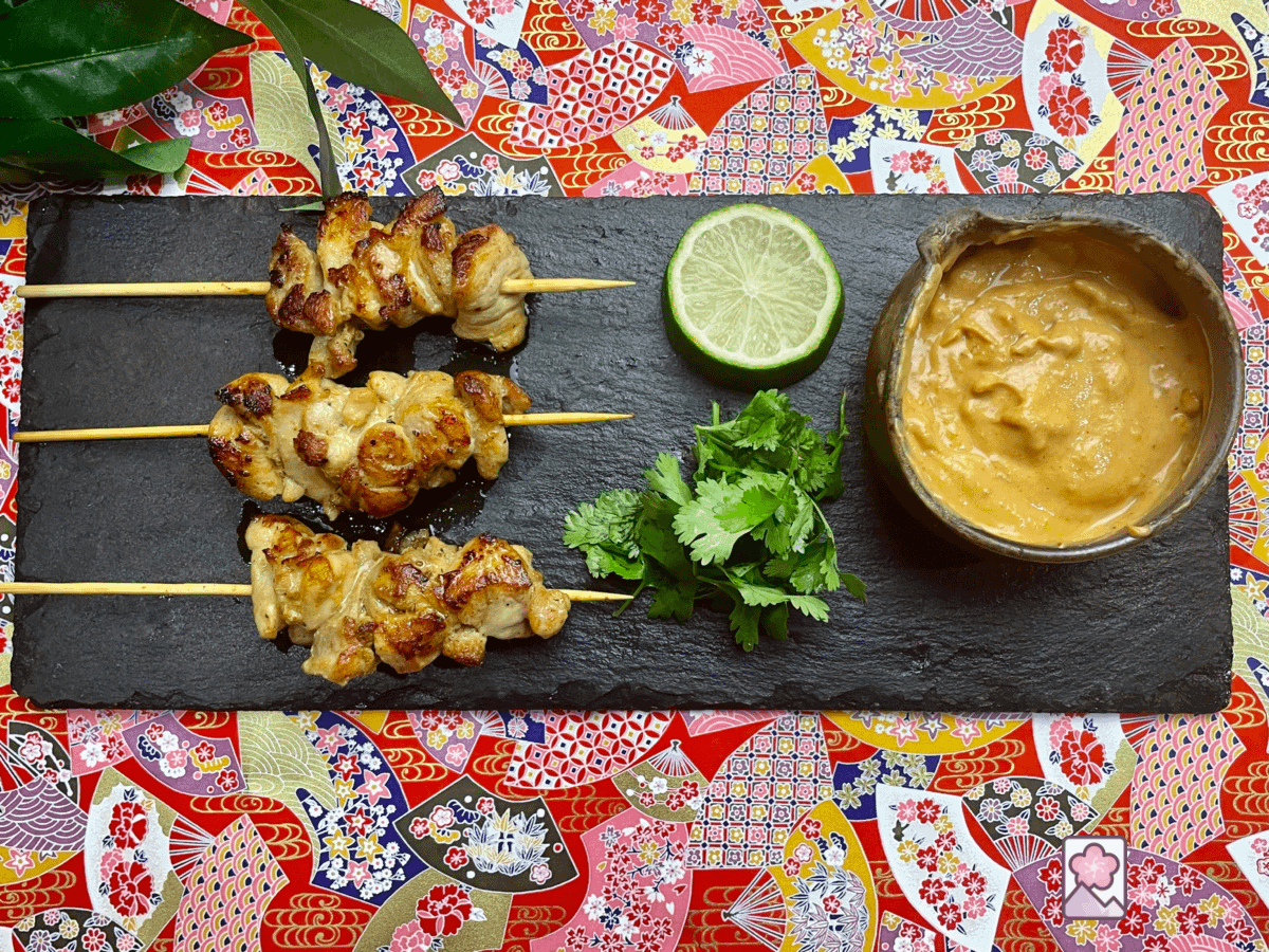 Three succulent chicken skewers served on a rectangular slate plate with peanut sauce, cilantro and fresh lime