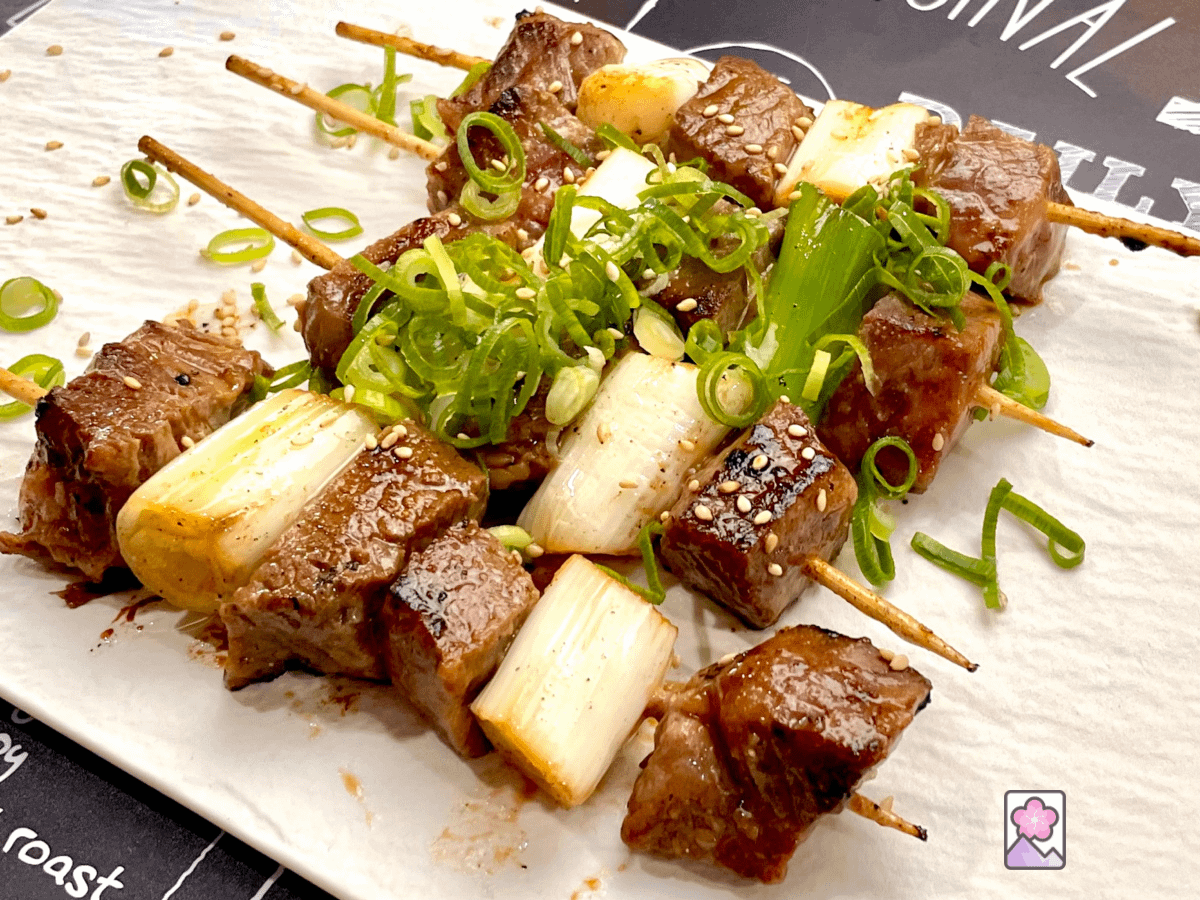 Four Miso Marbled Beef Skewers, alternating between tender Wagyu and cooked leeks. Are laid out on a rectangular ceramic plate.