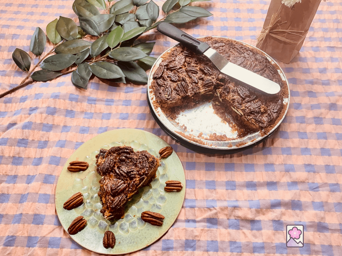 A slice of pecan pie on a small plate surrounded by pecans. There is a pie plate in the background with an offset spatula