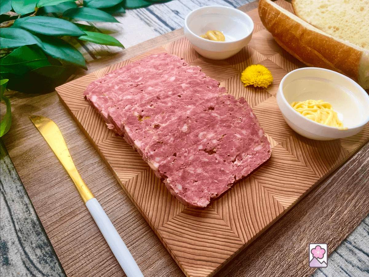 Homemade bologna, on a patterned wooden cutting board, with mustard, mayonnaise and a hard roll laid out for serving