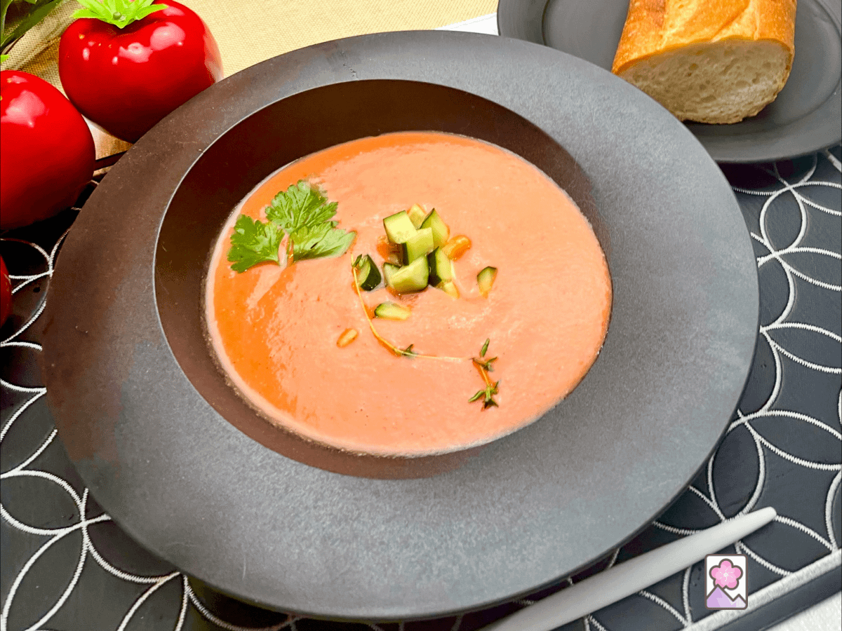 Chilled Gazpacho topped with chopped cucumbers, Italian parsley and thyme, served in a bowl with a piece of hard bread in the background