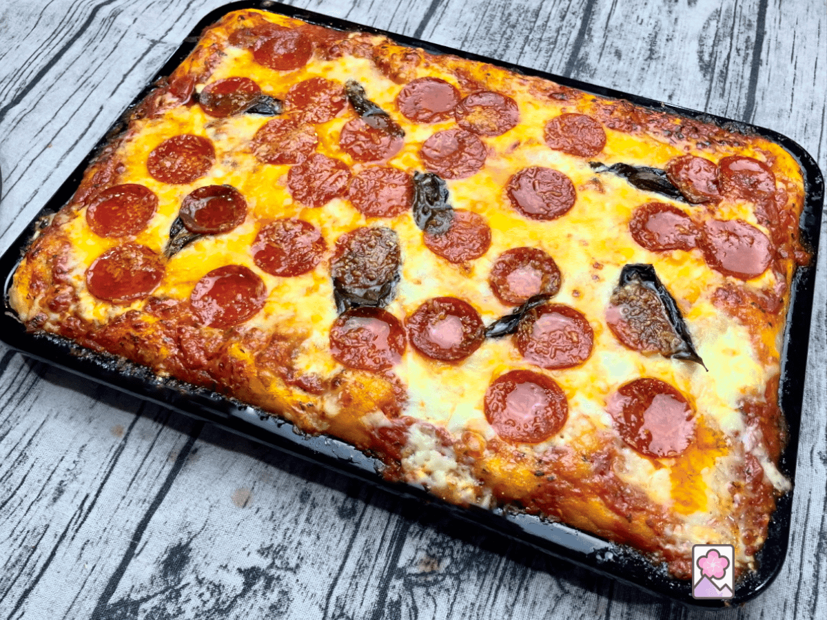 A Sicilian Pepperoni Pizza in a tray smothered with tomato sauce and loads of gooey cheese, to—Ed with fresh basil leaves and pepperoni slices