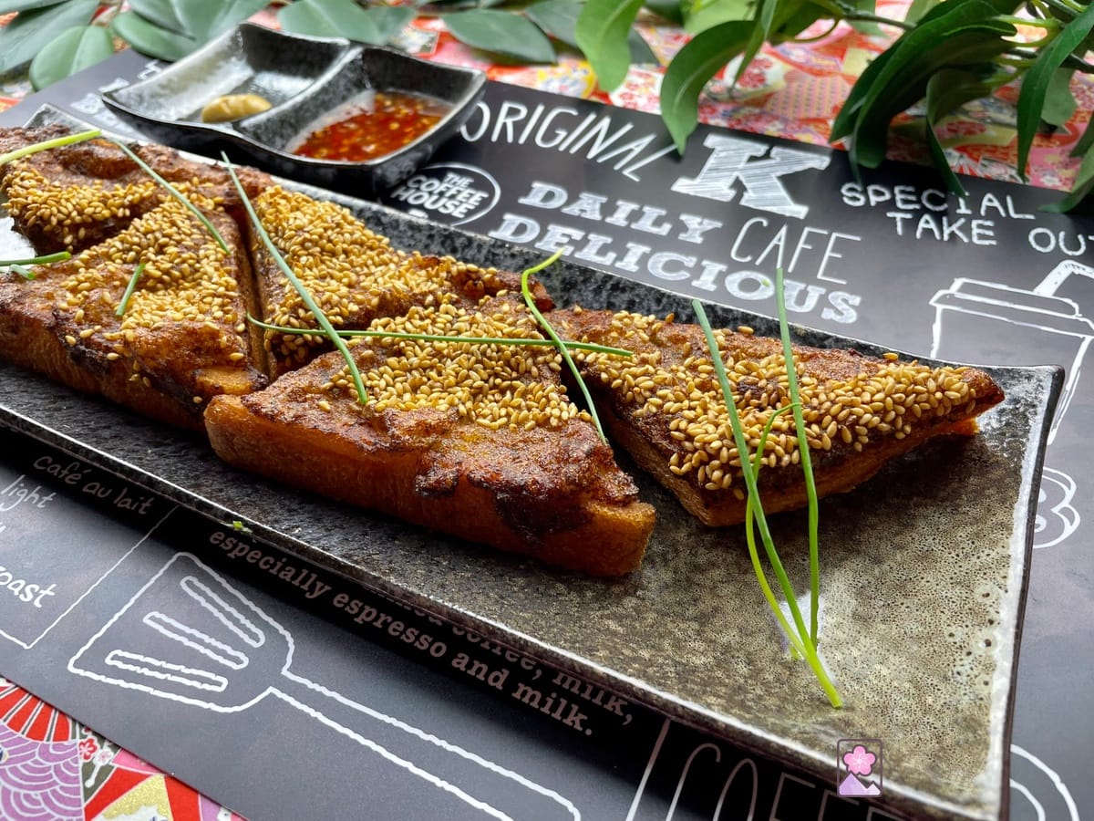 Shrimp Toast laid out on a rectangular ceramic plate topped with onion grass. There is a ceramic 2 compartment dish with sweet chili sauce and mustard behind it.