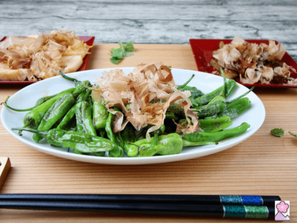 Sir Fried Shishito Peppers Served on a wood tray with other veggies behind it.