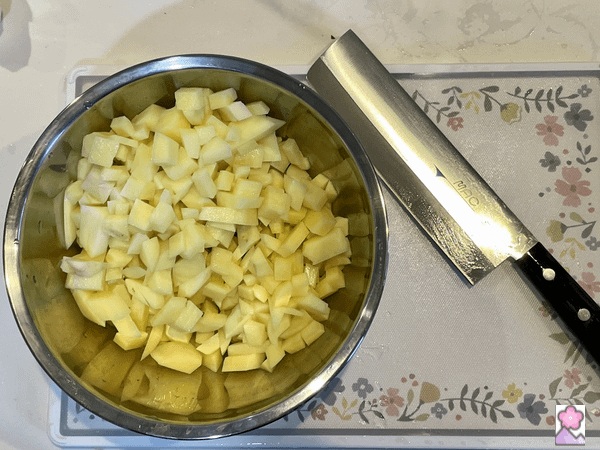 Washed, peeled and diced potatoes In a metal bowl on top of a cutting board. There is a knife to the right of the bowl.