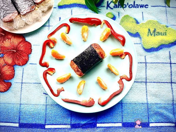 Spam Musubi on a plate surrounded by orange slices and red pepper slices. It’s on a plate on top of a cloth with a Hawaiian Islands map with another plate behind it that has several more Musubi wrapped in plastic wrap.