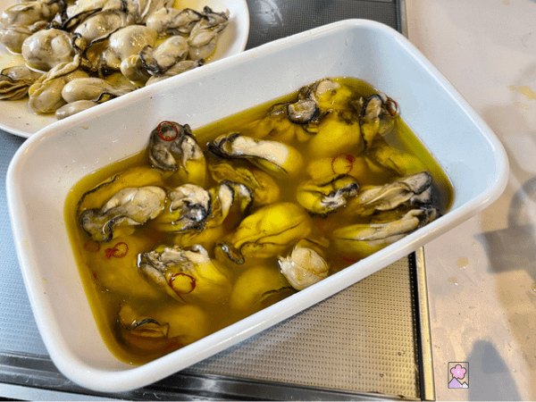 Oysters in the pan covered by marinade. There is a plate of cooked oysters in the background.