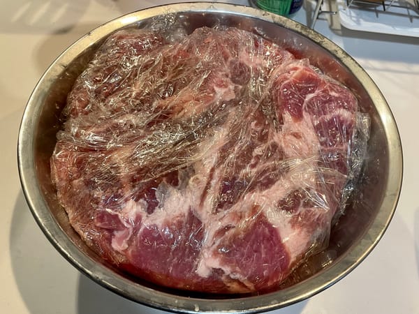 Pork shoulder after marinating in rub for 24 hours still wrapped in plastic in a large metal bowl