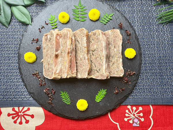 Five slices of Prime Rib Terrine wrapped in bacon from directly above. It’s on a black slate plate surrounded by flowers and leaves.