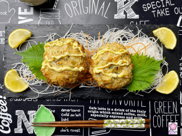 two Shellfish Cakes with Karasumi mayonnaise on top. THe cakes are on a garnish of daikon, carrot and shiso. The are lemon wedges on the plate.