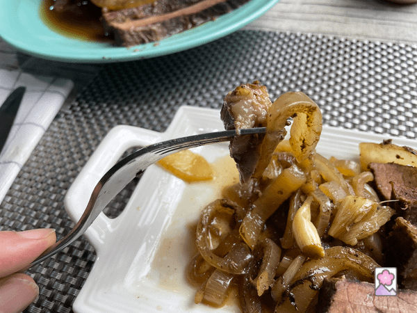 A piece of brisket on a fork smothered in cooked onions. The brisket is on an individual tray with a serving dish in the background.