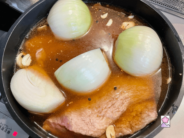 A brisket is submerged in a mix of water, consommé and spices. A quarter onion and garlic slices are also floating on top.