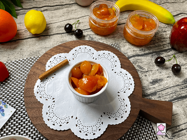 Kumquats in Syrup in a ramekin on a wood cutting board. There are a couple of glass containers of kumquats in syrup in the background along with fresh fruit