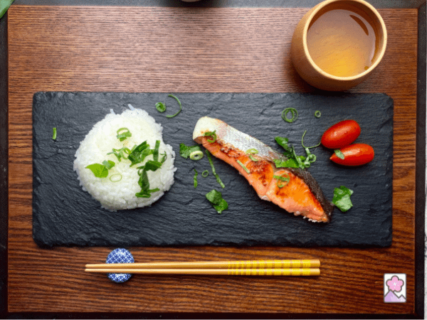 Koji Salmon with white rice, green tea, cherry tomatoes and sprinkled with fresh herbs. Served on a wooden tray with chopsticks.