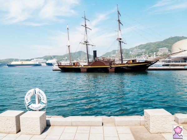 A 3 masted ship sits along a dock viewed from a wooden pier