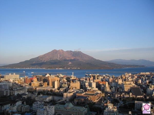 A city in the foreground. Behind it is a bay and across the bay is a large volcano.