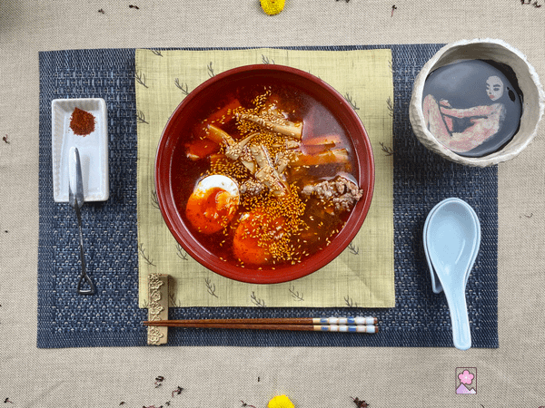 Tantanmen in a bowl with a spoon and chopsticks to the right and some chili powder to the left.