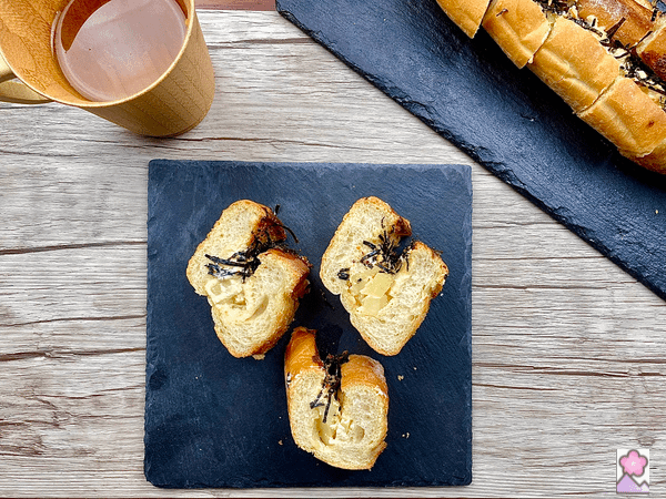 Mentaiko Potato Pan 3 slices on a square slate tray with another sliced baguette on a rectangular slate tray. There is a wooden cup of tea behind the square tray.